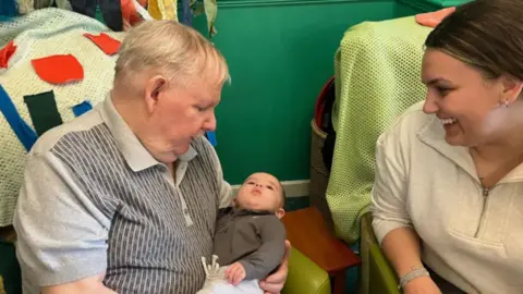 A care home resident holds a baby in his arms while his mother smiles sitting next to them.