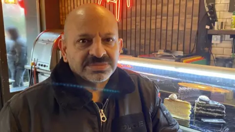 A man with a bald head and beard in front of a glass cabinet with cakes in it. He wears a zipped up black jacket.