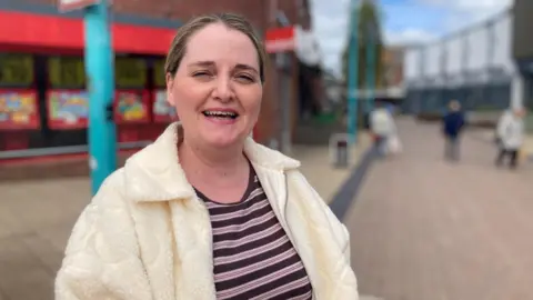 A woman, with blonde highlighted hair, smiles. She is in a stripey top and cream fluffy jacket. There are shops behind her.