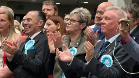 Smartly-dressed men and women with Reform UK rosettes pinned on their jackets. They are clapping and smiling. There is a hand holding a phone taking a picture on the far right.