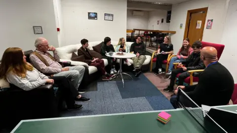 A group of ten teenagers and parents are sitting in a semi-circle in a room at a youth club. The furniture includes sofas and armchairs. In the foreground there is a table tennis table.