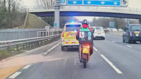 A grainy screenshot from some dashcam footage of a motorcycle rider in front, in the left hand lane of a motorway. There's a police car directly in front of him with its blue lights flashing.