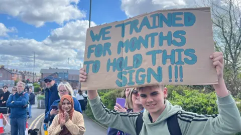 Teenage boy holds up a cardboard sign saying "We trained for months to hold this sign!". Dozens of spectators line the route next to him, applauding and watching runners.