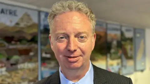 Alex Allinson, who has short, fair, wavy hair, is wearing a dark pinstriped suit jacket and light blue shirt.