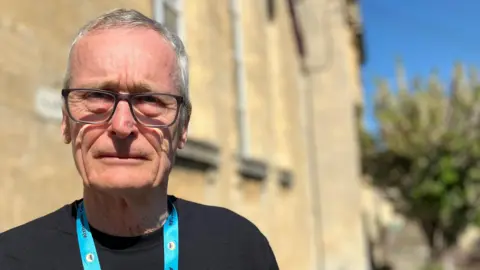 A man with glasses and grey hair is looking directly at the camera. He is wearing a blue 'Alzheimer's café' lanyard and a black T-shirt.