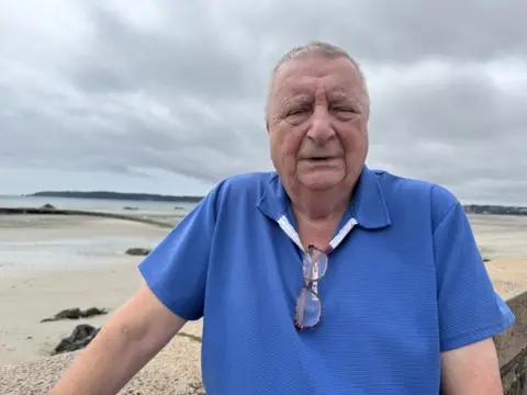 A man stands outdoors near a beach, wearing a blue short-sleeved shirt with glasses hanging from the collar. The background features a sandy beach, rocks, and an overcast sky.