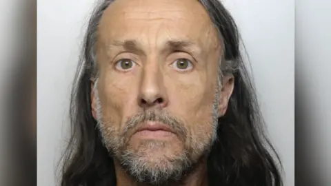 Dyfed-Powys Police Mugshot of Richard Jones who has long, shoulder-length grey hair and a grey beard and moustache and is looking straight at camera with a neutral expression.