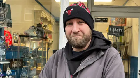 Federica Bedendo/BBC Paul Johnston is standing outside a shop and is wearing a black and red hat, a black hoodie and grey coat. He has blue eyes and a beard.