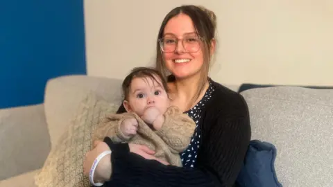 Chloe with her brown hair tied back, looks into the camera smiling. She is wearing a black cardigan over a black and white patterned dress. She is cuddling her six-month-old baby who has her fist in her month and is wearing a knitted, beige cardigan.