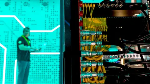 A man standing against a bright green background in a data centre - in the foreground is the back of a large computer with lots of yellow and black wires.