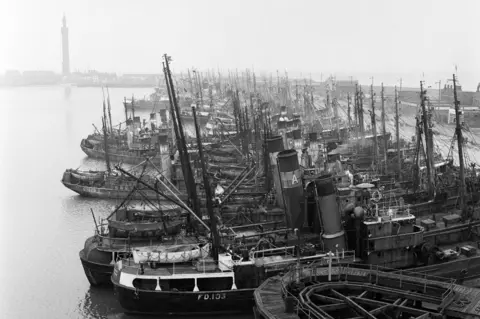 Getty Images A black and white photo of dozens of trawlers and fishing boats docked closely together in a calm-looking dock. A tall tower can be seen in the background and low buildings can be seen to the right.