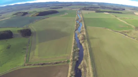 Lifewader A river flows in a fairly straight line between two high embankments. On either side are large agricultural fields full of crops 