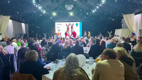 St Elizabeth Hospice A crowded room captured from behind showing hundreds of people sat at tables as an auction takes place. Some hare sculptures can be seen on stage and on the screen. 
