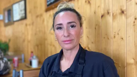 A woman has blonde hair tied in a bun and is wearing a navy blue shirt with a navy blue apron. She is sitting on a chair next to a wall with wooden panelling. There are tables with condiments on them behind. 