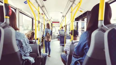 A group of people sitting on a bus. The picture of the bus has been taken from the inside and has been taken from the back of the vehicle. There are about six people all with theirs backs to the camera, including one woman who is standing up, while the others are all sitting down. On the inside, the bus has yellow railings for passengers to hold on to.
