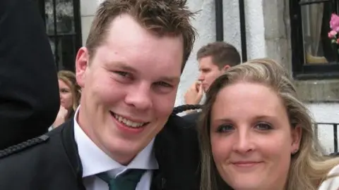 Felicity and James Hester are a couple in their thirties. They are both smartly dressed with James wearing a black jacket and tie. Only Felicity's head is visible but she has long blonde hair. He has his arm around her and they are both smiling.