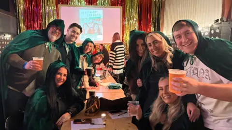 A group of men and women in dark green cloaks pose either side of a long wooden table holding drinks and smiling.