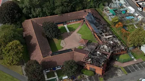 An aerial photo of the fire-damaged office. The entire roof of the square-shaped brick building has been destroyed with blackened beams visible. There is a courtyard in the middle of the building with a paved square surrounded by grass. There are trees surrounding the building and a row of allotments on the right.