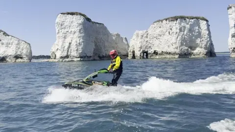 A jet skier in a yellow drysuit and red helmet rides a stand-up jet ski through calm, bright blue waters, passing large white chalk sea stack cliffs under a clear sky.