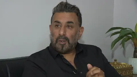 Shabaz Hussain sits on a sofa with his hands clasped. He wears a black shirt and has short black hair and a short black and gray beard. 