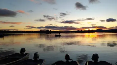 Nikki Forde Rowboats rest on a still lake as the sun sets behind distant hills.
