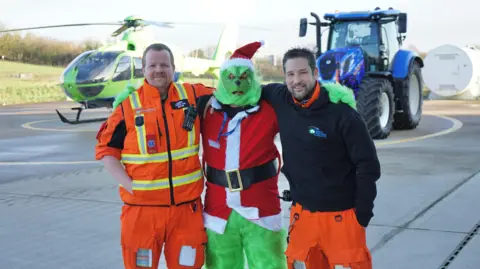 Handout Mr Gibson posing for a photo with two members of the Great Western Air Ambulance. He is dressed as the Grinch wearing a Santa outfit and a Christmas hat, standing with his arms around two other men, who are wearing hi-vis orange uniforms. In the background there is a green helicopter and a blue tractor parked on a helipad. 