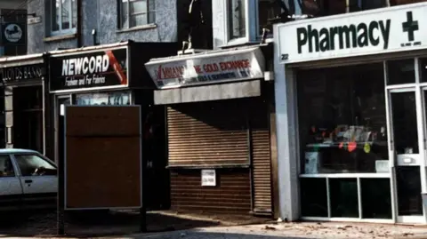 Hampshire Constabulary Archive picture of jewellery shop with shutters down, in between a pharmacy and fabric shop on Shirley Road