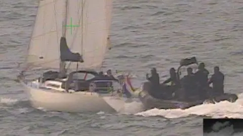 National Crime Agency This is an image captured by CCTV. On the left of the picture is the yacht used by the people smugglers with its sails up. On the right a rigid inflatable boat with border force officers dressed in black preparing to board the vessel. 