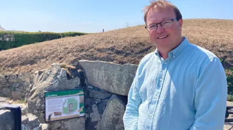 Macey Turner/BBC Martyn Dorey is wearing a blue shirt and he is standing beside the entrance to the burial chamber which is built of large blocks of stone covered by soil with parched grass on the top.