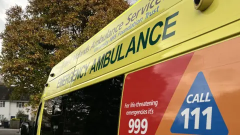 A close-up shot of the side of an ambulance. At the top it says West Midlands Ambulance Service University NHS Foundation Trust. Below that in larger lettering it says emergency ambulance, and at the bottom in white lettering against a red background it says for life-threatening emergencies it's 999