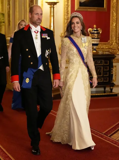 Aaron Chown / Pool El Príncipe de Gales está en el tradicional código de vestimenta de corbata blanca y está decorado con sus órdenes reales y su regalía. Su chaqueta tiene solapas rojas. La princesa de Gales usa un vestido de alta costura del diseñador británico Phillipa Lepley con un abrigo de noche de encaje de chantilly dorado de cuerpo completo sobre un vestido de crepe de seda. Ella lleva la tiara de nudos de un amante.