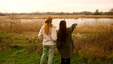 Two women looking out over a wetland reserve with white reeds in the foreground. The woman on the left has red hair with a cream jacket and black t-shirt with light green trousers. The woman on the right, who is pointing into the distance, has long brown hair and is wearing a green full-length coat with black trousers