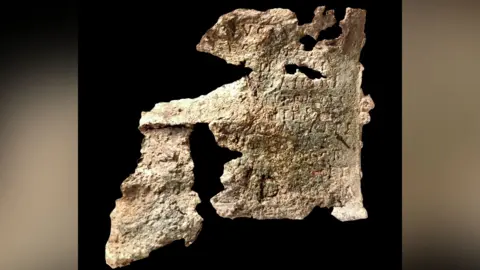 A damaged old bronze plaque on a dark background. Words etched in the metal can just be made out but the plaque has may holes in it.