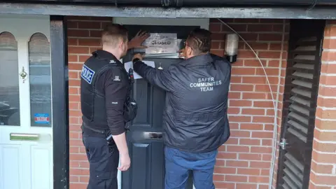 A police officer and member of the Safer Communities Team place a sign which reads "closed" on a dark grey front door surrounded by red brick.