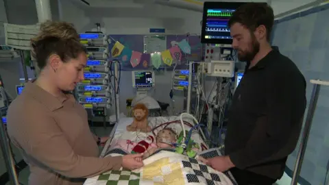 Hollie and Jordan stand either side of a hospital bed, with Penelope in the bed connected to many wires and equipment