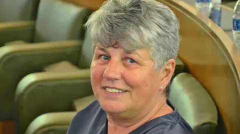 Robbie MacDonald/LDRS West Lancashire Borough Council leader Yvonne Gagen with short grey hair, blue eyes, wearing a grey top smiling as she sits on a green leather seat