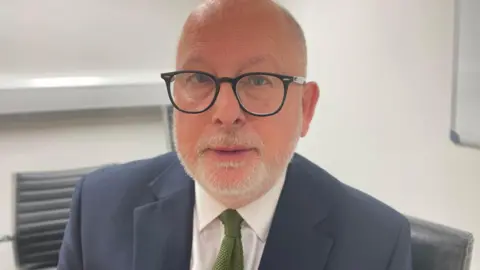 Steve Yemm is looking directly at the camera. He has a shaved head and is wearing black rimmed circular glasses. He has a navy suit jacket with a white shirt and a green tie.