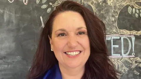 Grace Wood/BBC A woman with long brown hair smiles at the camera. She is standing in front of a blackboard