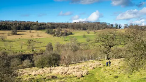 Tim Graham/Getty Images A beautiful Cotswolds valley landscape in the sunshine with a blue sky with some clouds above it. A house surrounded by trees can be seen on one slope, as a pair of walkers make their way through farmland on the other.