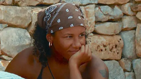 BBC/Twofour Lailah on I Kissed A Girl. Lailah has curly brown hair to her shoulders and wears a brown bandana. She has brown eyes and rests her chin on her hand while looking off camera. She's pictured outside at the Italian villa wearing a black bikini. 