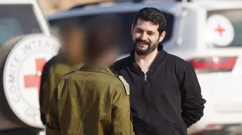 IDF Ziv Berman speaks to two IDF soldiers who have their faces blurred, with Red Cross vehicles in the background.