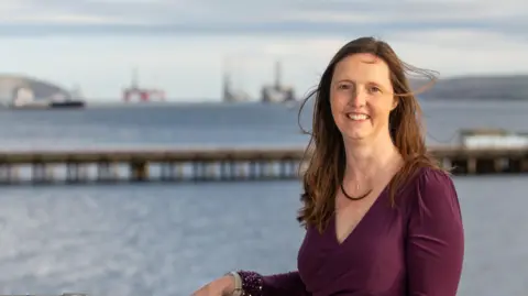 Port of Cromarty Firth Joanne has long brown hair and is wearing a dark top. She is smiling and pictured with the Cromarty Firth in the background. It is a sunny day.