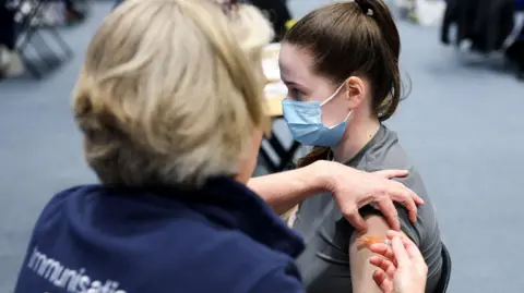 A woman in a blue face mask is being injected by another woman. The person being injected has long, brown hair in a ponytail and is wearing a grey shirt. The needle is mainly obscured by the other person's hand, but it has an orange cap. The person with the needle has mid-length blonde hair and the word immunisation printed in white on the back of her blue jacket.