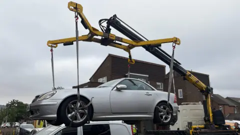 Silver coupe suspended by a crane during a vehicle recovery operation, being lifted over a white van in a fenced area surrounded by brick buildings.