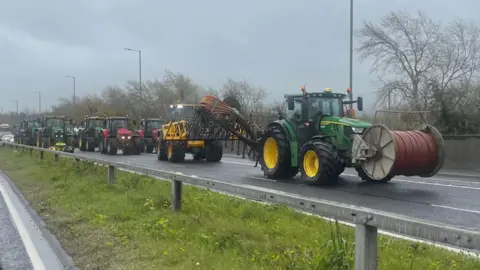 A number of tractors drive along a road behind one another, with some traffic seen in the distance behind them. The tractors are a range of colours and some have equipment attached.