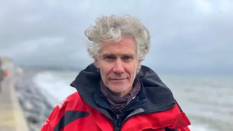 A man stands on the seafront at Torcross, Devon. There are waves crashing against the sea defences in the background. He is wearing a red waterproof jacket. 