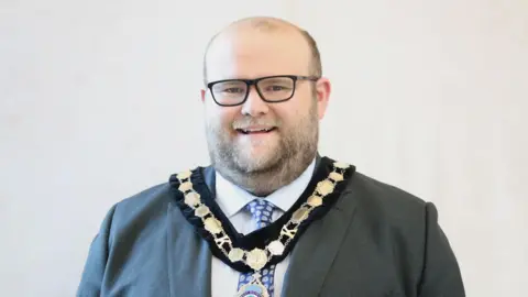 A portrait shot of Councillor George Connolly. He has a partially bald head and a light brown beard. He is wearing a ceremonial gold chain over his grey suit.