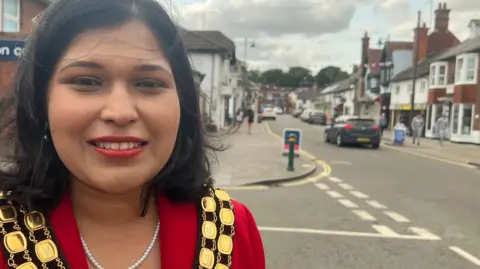 Image of a woman wearing the chains of office of a mayor. In the background can be seen a high street with shops, traffic lights and cars parked.