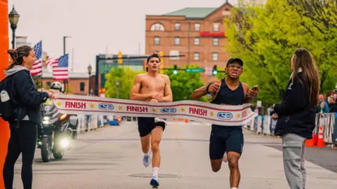 The finish line of a race, one man with no top behind the ribbon and another runner just going through the ribbon.