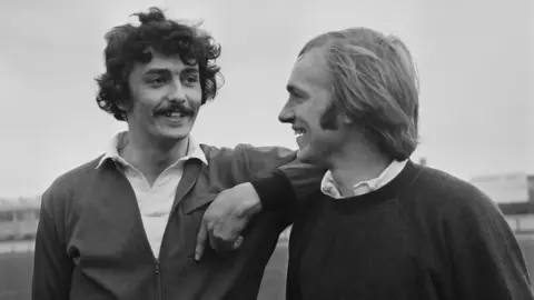 Two men stand side-by-side. The man on the left has black curly hair and a moustache. His elbow is resting on the shoulder of the man on the right who has fair hair and is wearing a jumper and open-necked shirt.